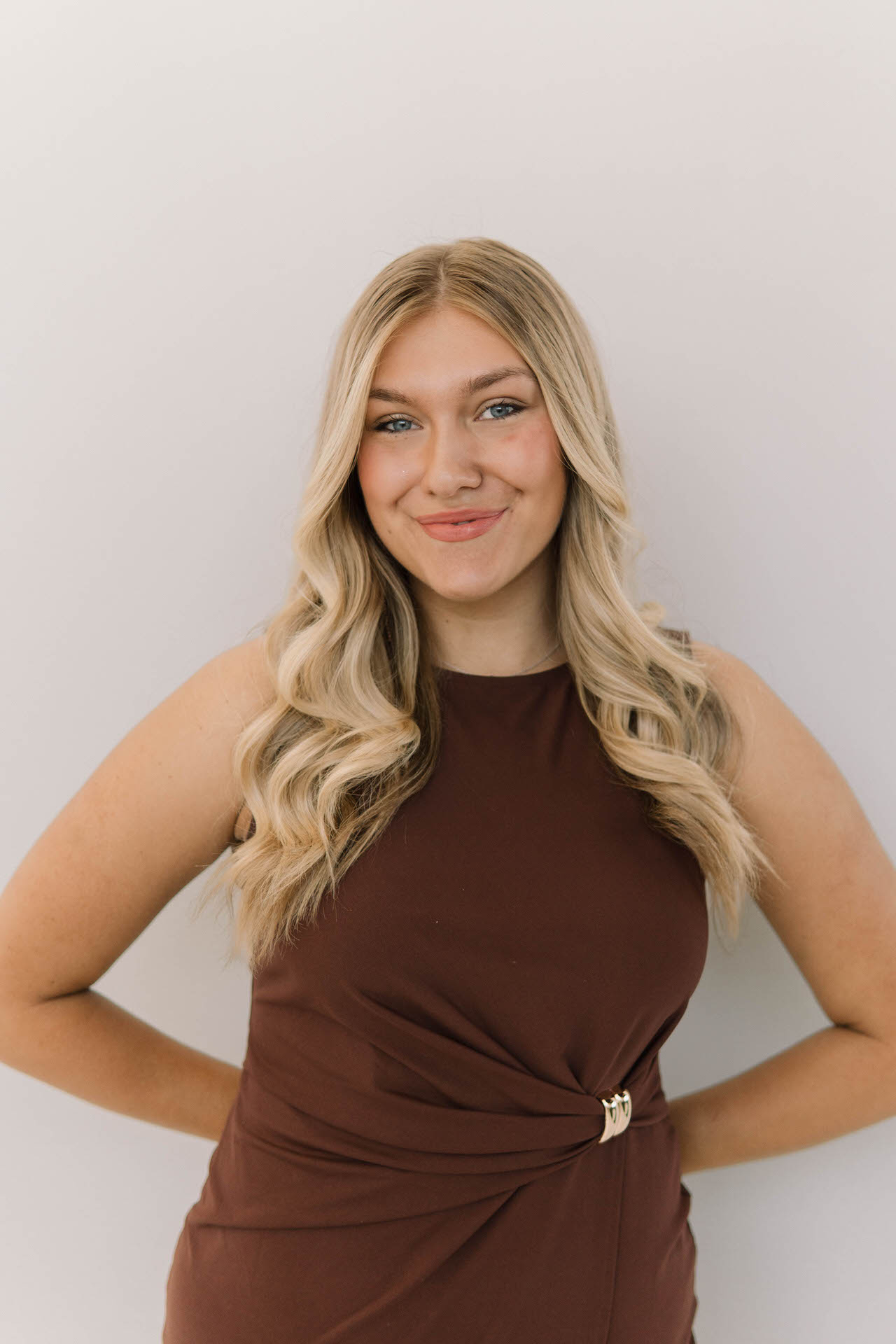 A young woman with long blonde wavy hair, wearing a sleeveless brown dress, stands against a plain light-colored background, smiling at the camera.