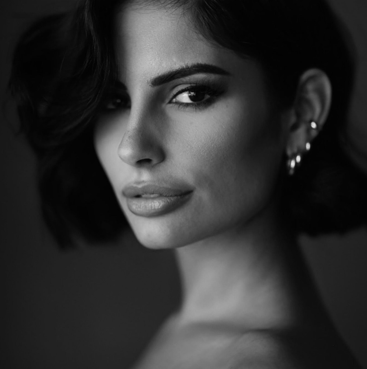 Black and white portrait of a woman with short, wavy dark hair, arched eyebrows, and multiple earrings, gazing confidently at the camera.
