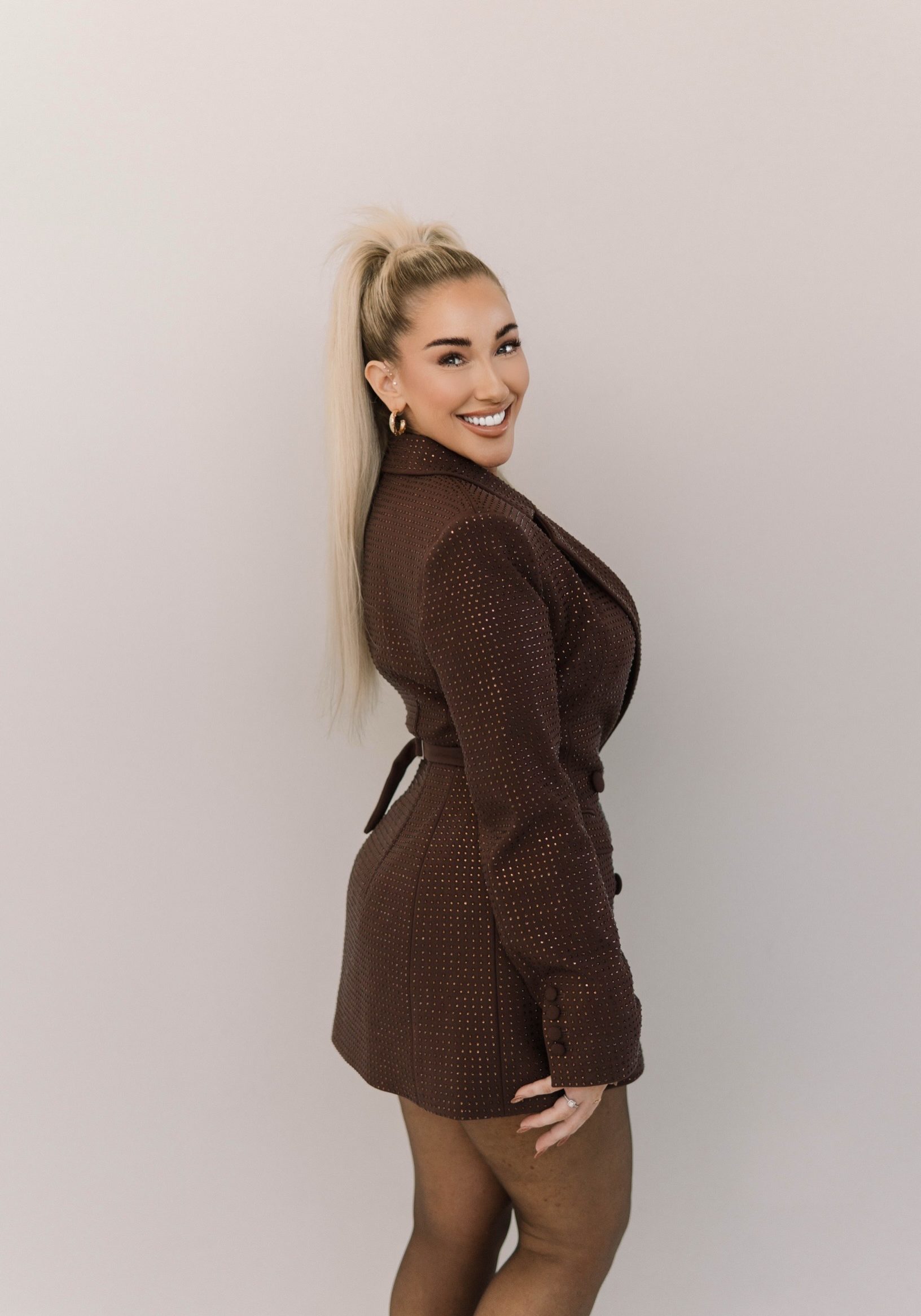 A woman with long blonde hair in a high ponytail wears a brown belted blazer dress and smiles while posing against a plain light background.
