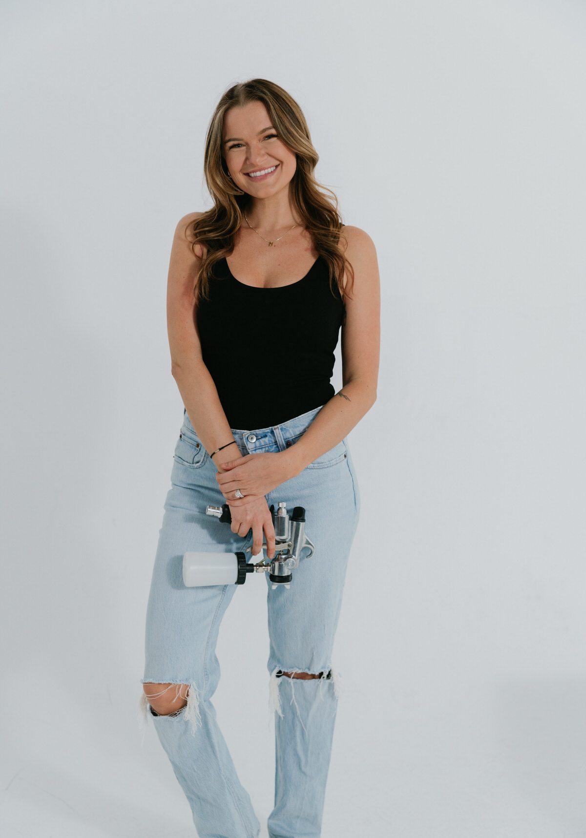 A woman in a black tank top and ripped light blue jeans stands smiling and holding a paint spray gun against a plain white background.