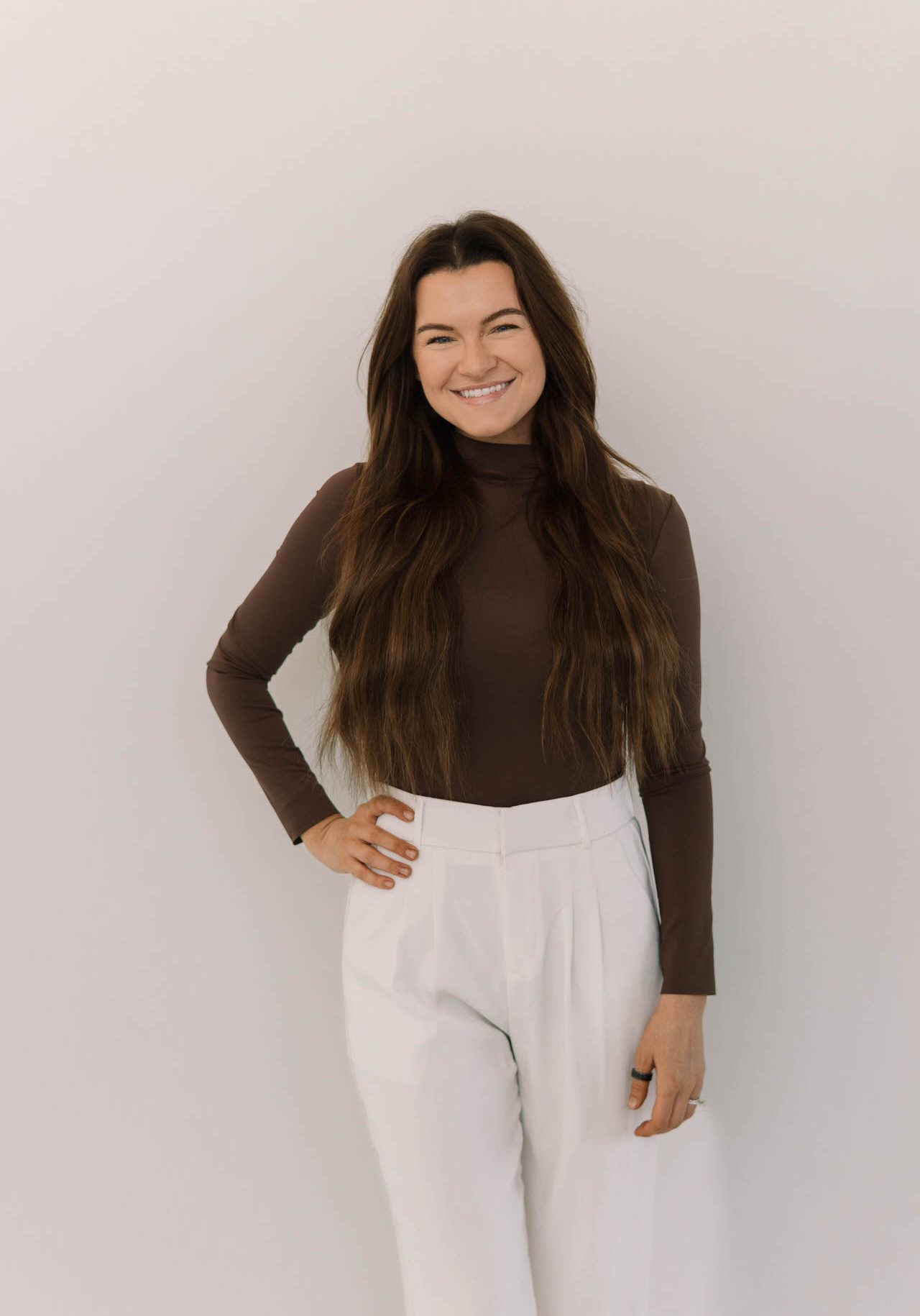A woman with long brown hair, wearing a brown turtleneck and white pants, stands against a plain light background, smiling with one hand on her hip.