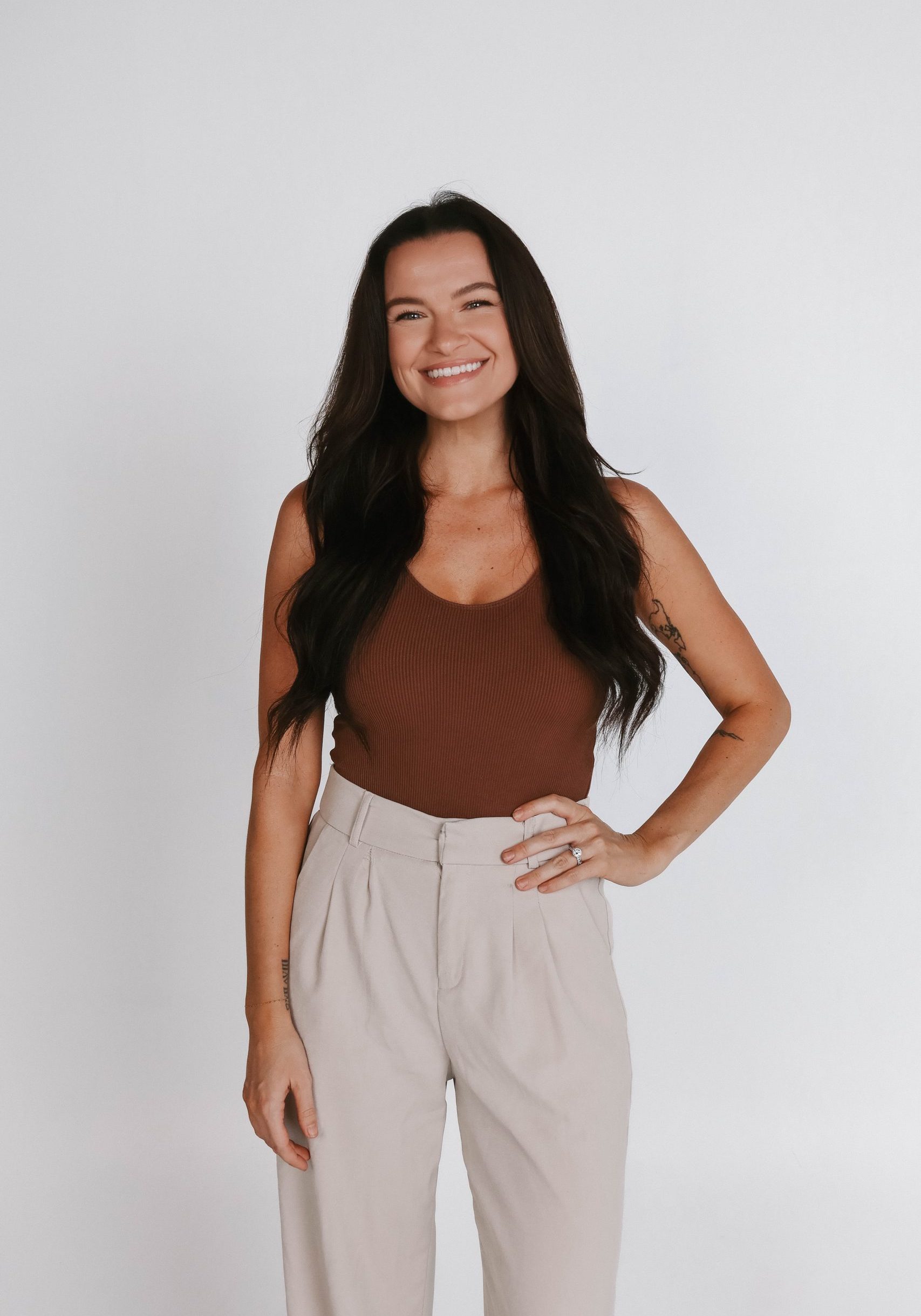 Woman with long dark hair, wearing a brown sleeveless top and beige high-waisted pants, stands against a plain white background, smiling with one hand on her hip.