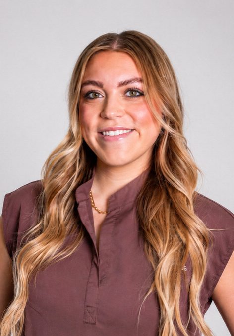 A woman with long, wavy blonde hair wearing a brown short-sleeve blouse stands against a plain light gray background, smiling at the camera.