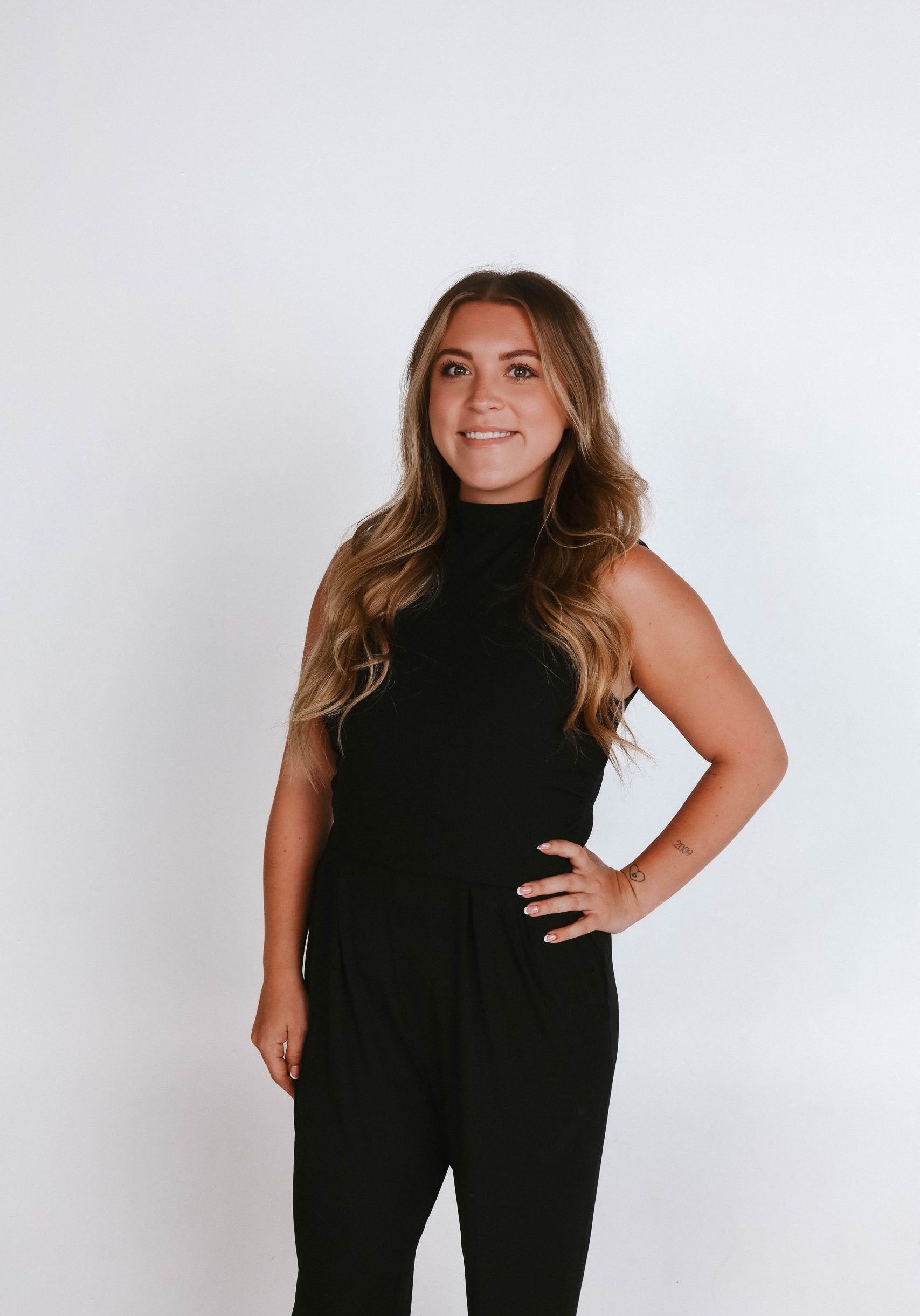 Woman with long wavy hair stands against a plain white background, wearing a sleeveless black top and pants, with one hand on her hip and a neutral expression.