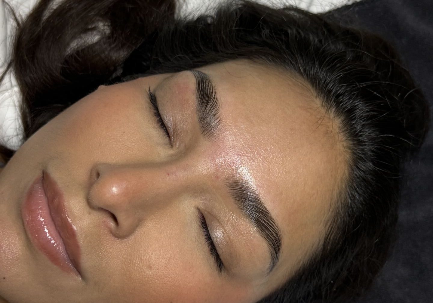 A woman with long dark hair and well-groomed eyebrows lies on a white surface with her eyes closed, resting her head on a dark cloth.