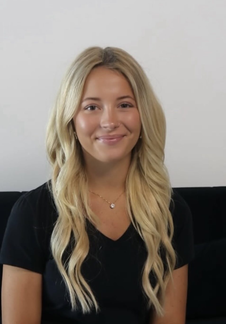 A woman with long blonde hair and a black shirt sits on a black sofa in front of a plain white wall, smiling at the camera.
