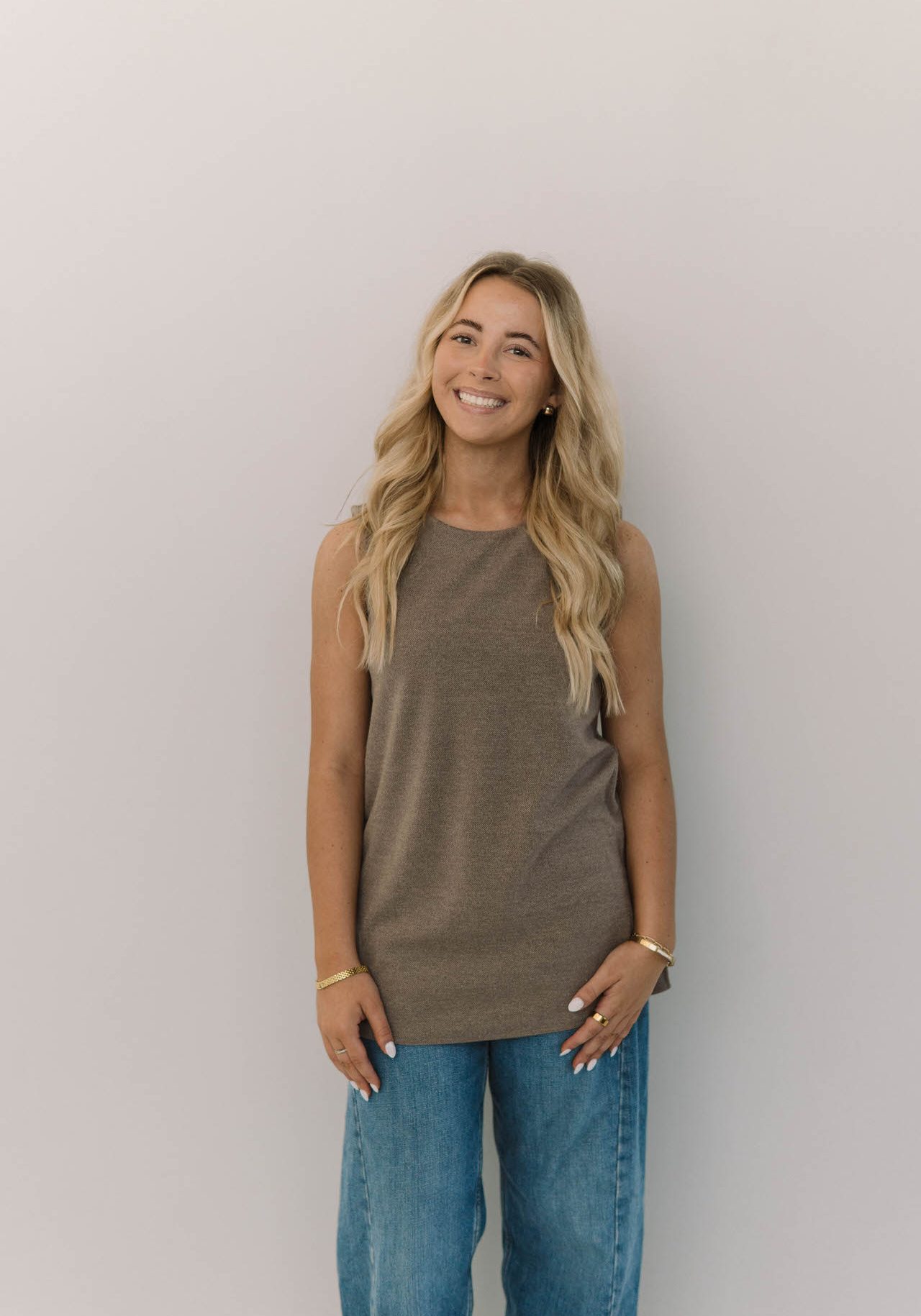 A woman with long blonde hair, wearing a sleeveless brown top and blue jeans, stands against a plain light-colored background, smiling at the camera.