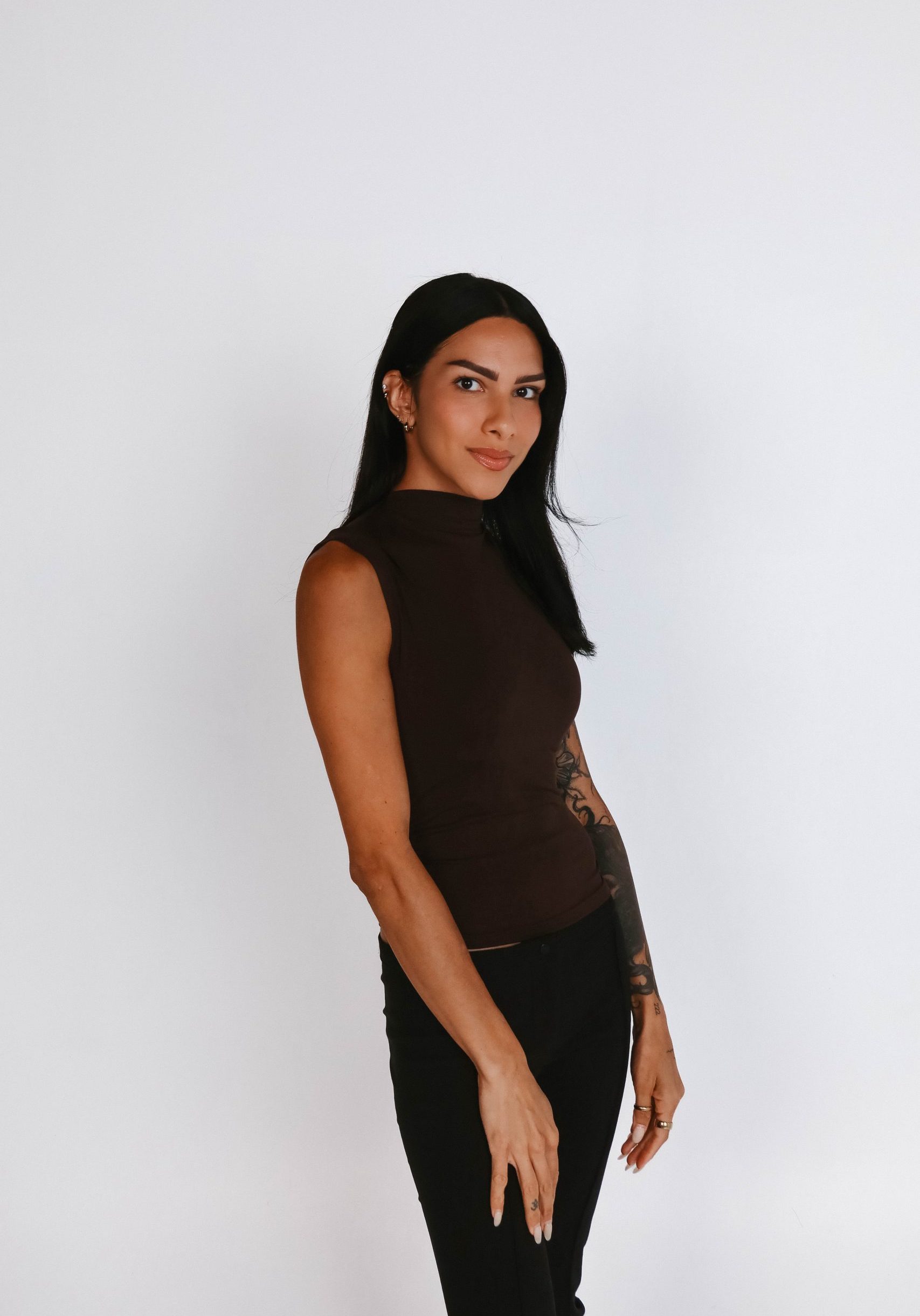 Person with long dark hair, wearing a sleeveless dark brown top and black pants, standing against a plain white background.