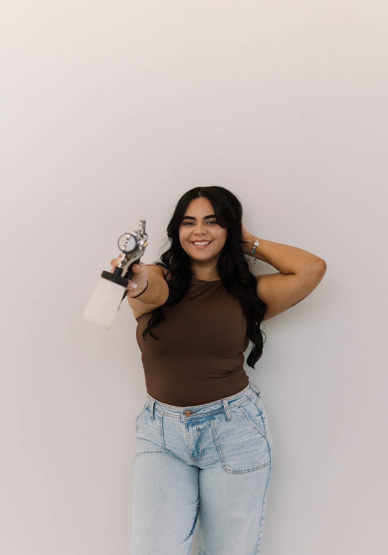 Woman with long dark hair in a brown sleeveless top and light blue jeans stands against a plain backdrop, smiling and holding an airbrush spray gun toward the camera.