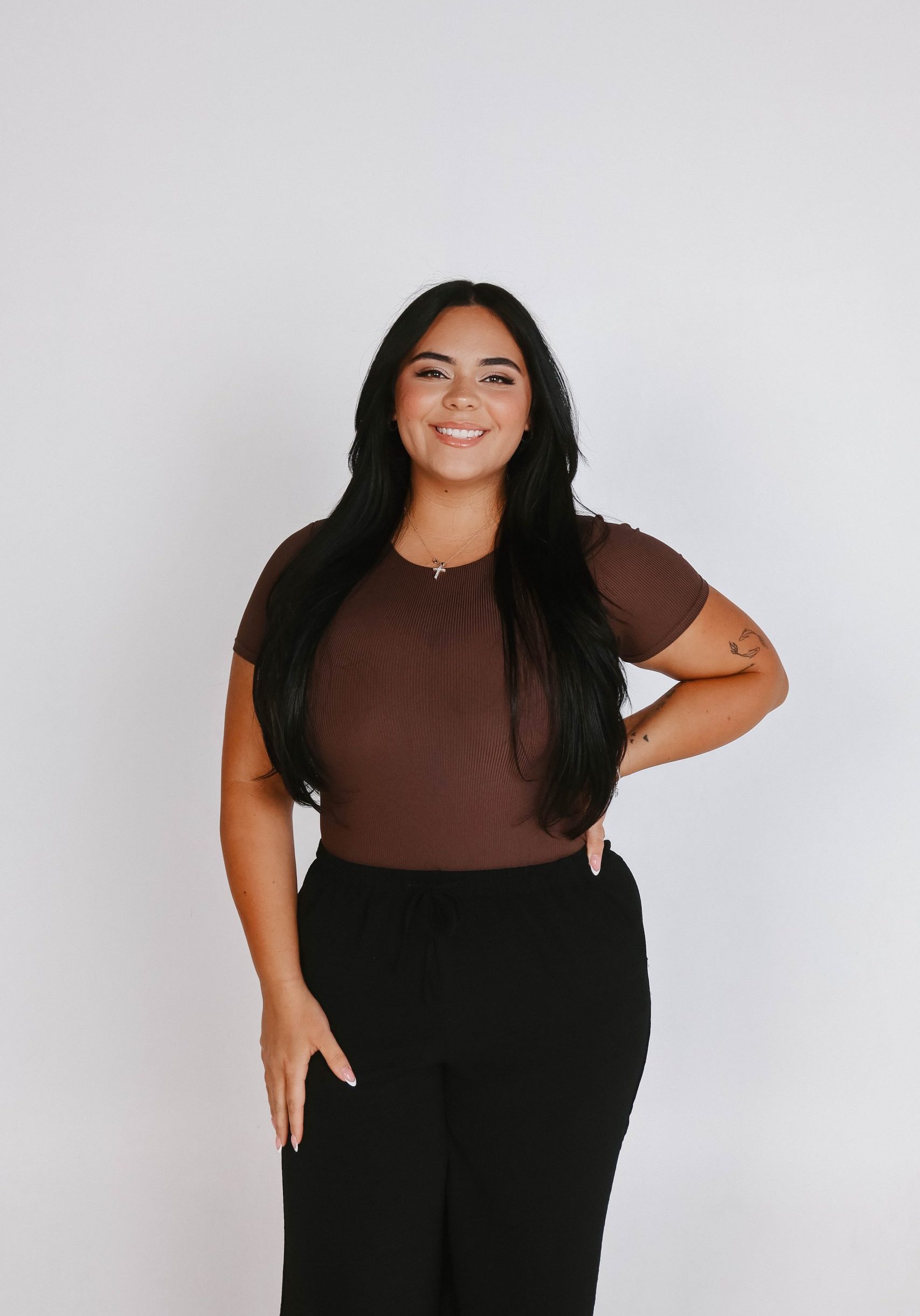 Woman with long dark hair wearing a brown top and black pants stands smiling against a plain white background.