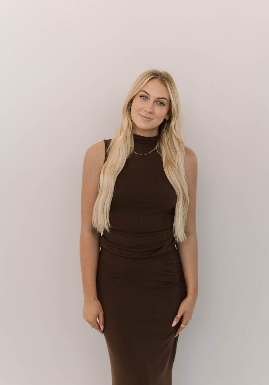 A woman with long blonde hair wearing a sleeveless brown dress stands against a plain light-colored background, looking at the camera.