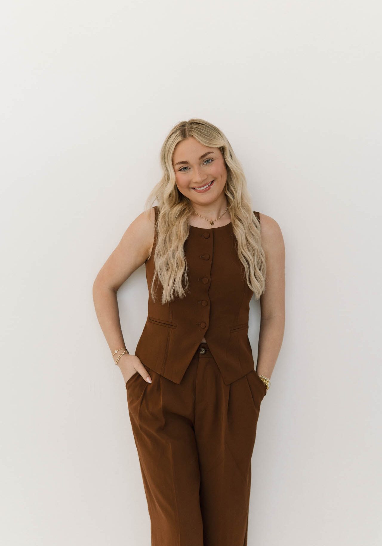 A woman with long blonde hair in a brown sleeveless vest and matching pants stands against a white background, smiling with hands in her pockets.