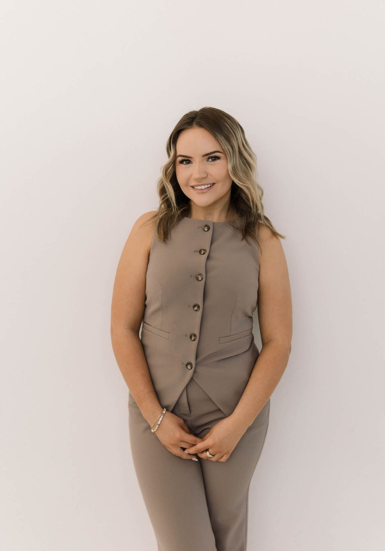 A woman with wavy, shoulder-length hair stands against a white background, wearing a beige sleeveless top and matching pants, smiling with her hands clasped in front.