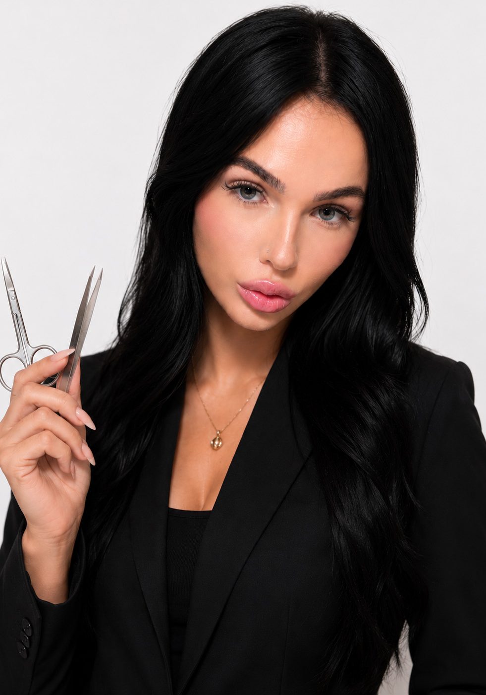 Woman with long black hair in a black blazer holds small scissors in her right hand against a plain light background.