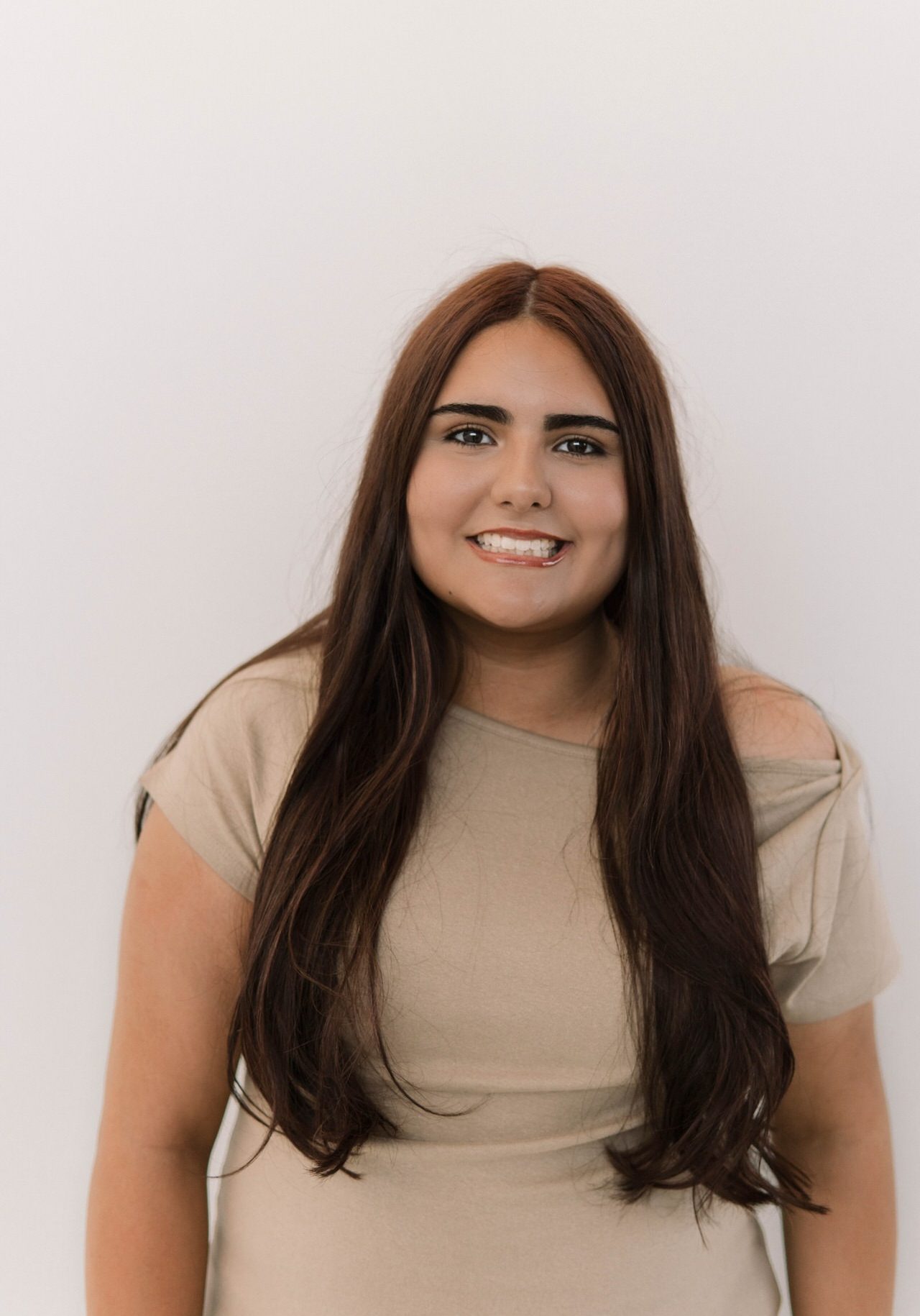 A young woman with long brown hair wearing a beige top stands against a plain white background, smiling at the camera.