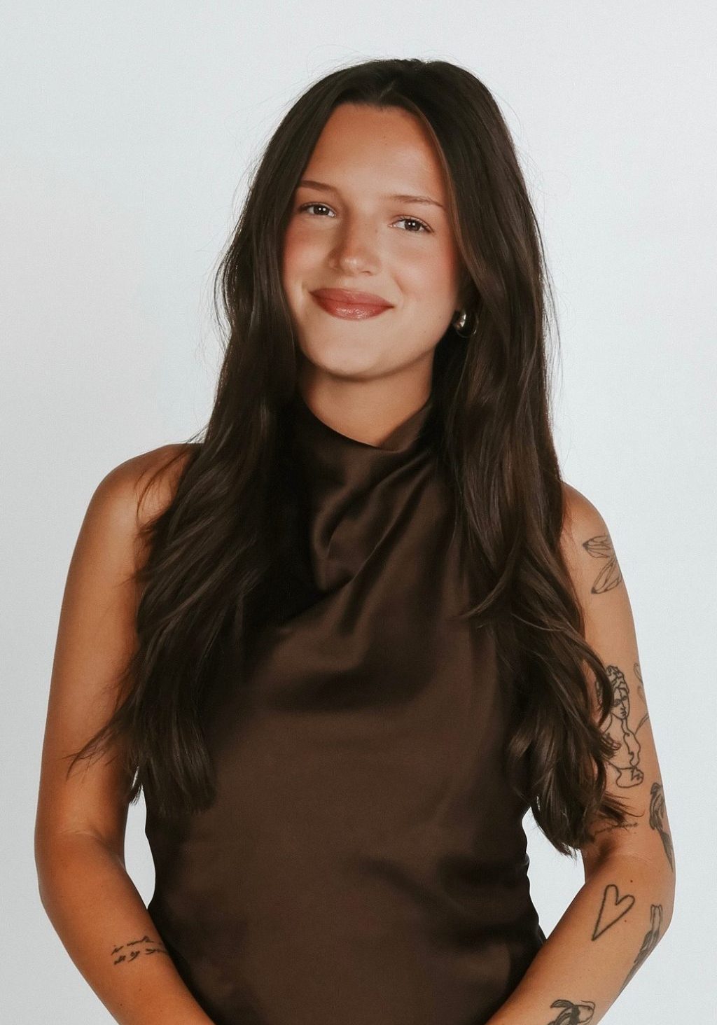 A woman with long brown hair, wearing a sleeveless brown top, stands smiling with her hands in front of her. She has visible tattoos on both arms and is posed against a plain light background.