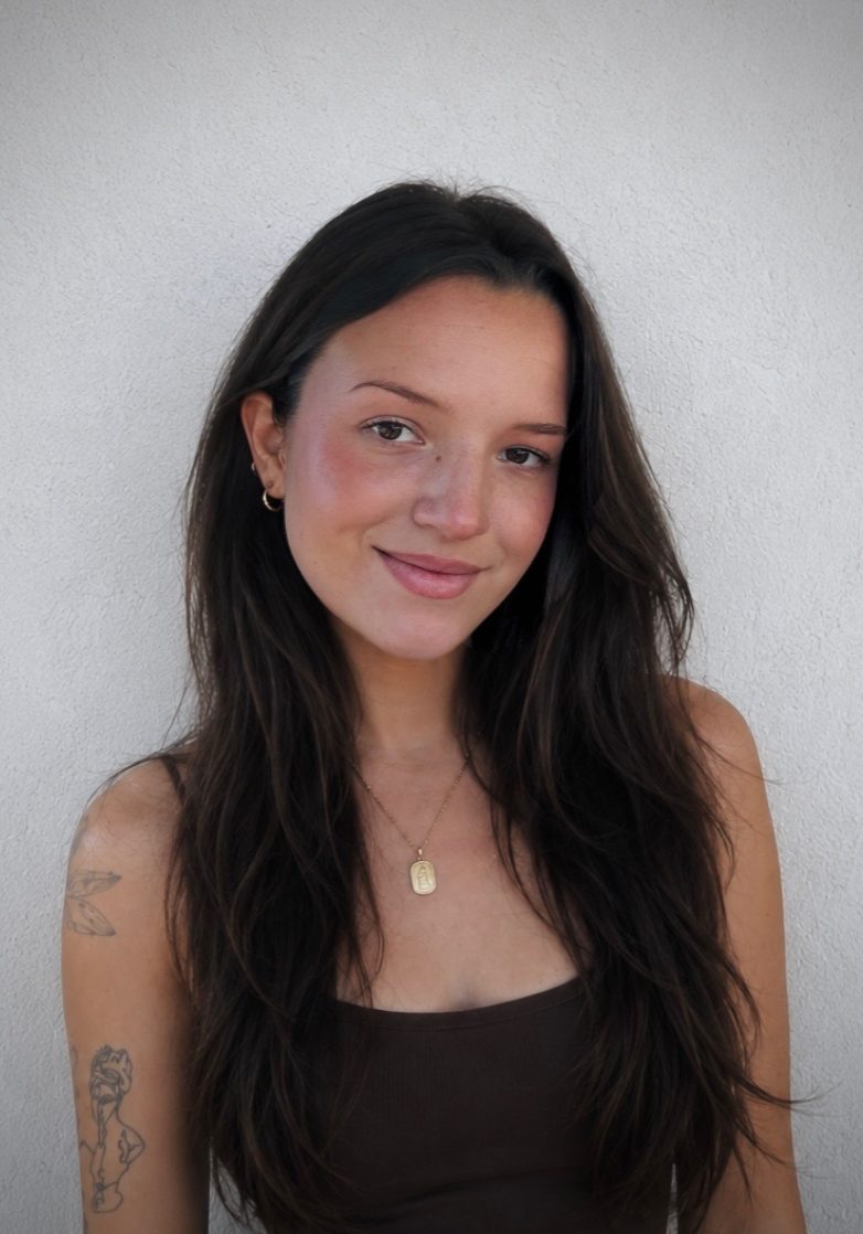 A woman with long brown hair and a brown tank top stands in front of a white wall, smiling slightly. She has a tattoo on her arm and wears a gold necklace and small earrings.
