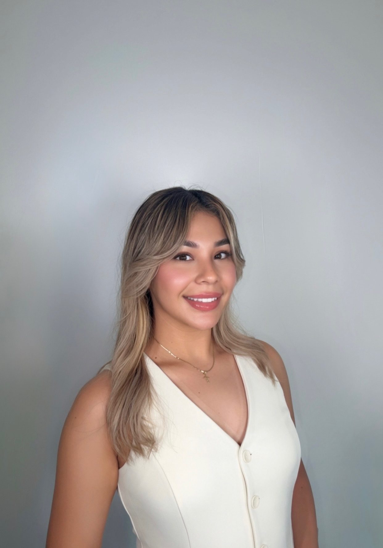 Woman with long, wavy blonde hair wearing a sleeveless white buttoned top poses and smiles in front of a plain light gray background.