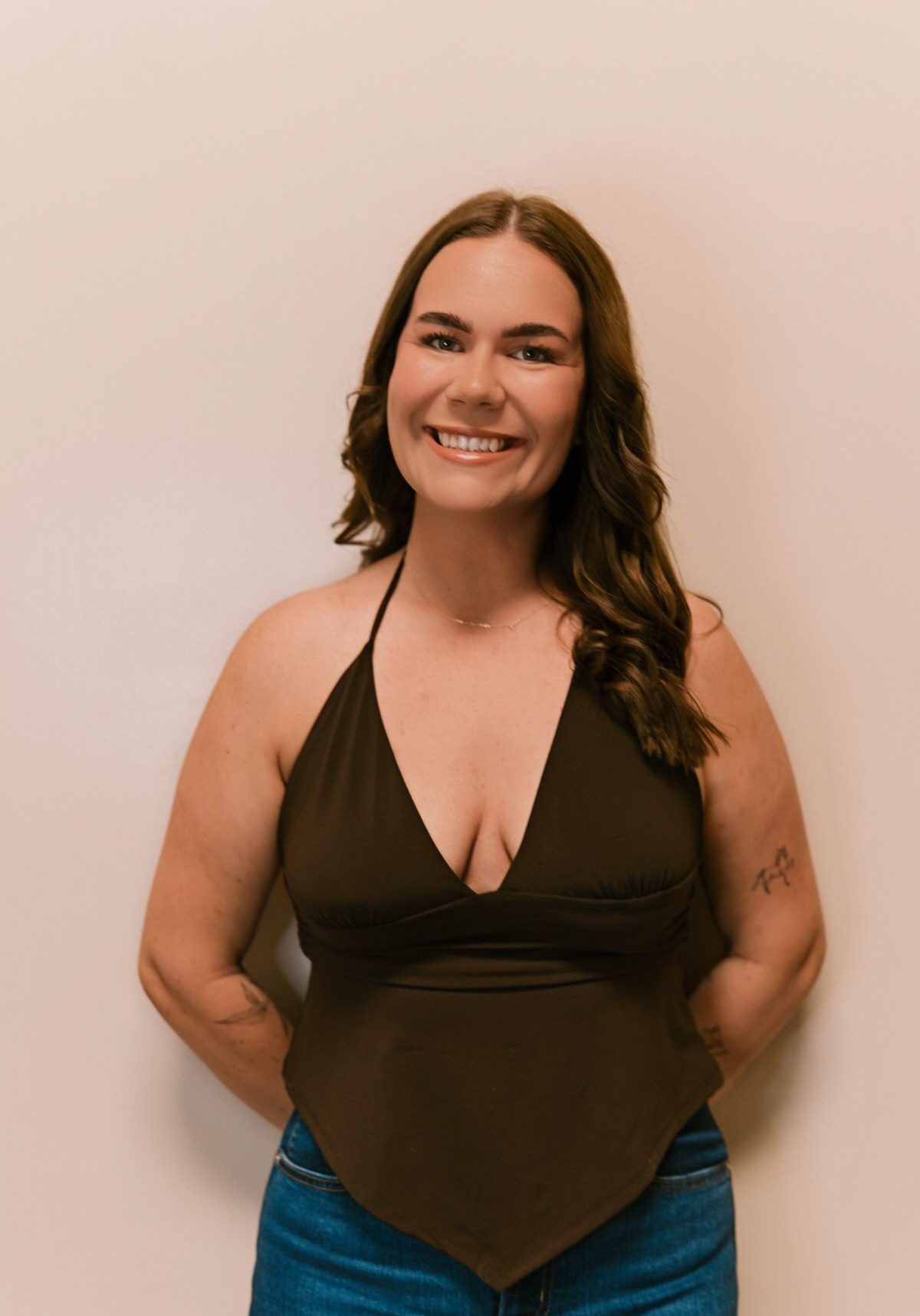 A woman with long brown hair wearing a dark halter top and jeans stands against a plain light-colored wall, smiling with her hands behind her back.