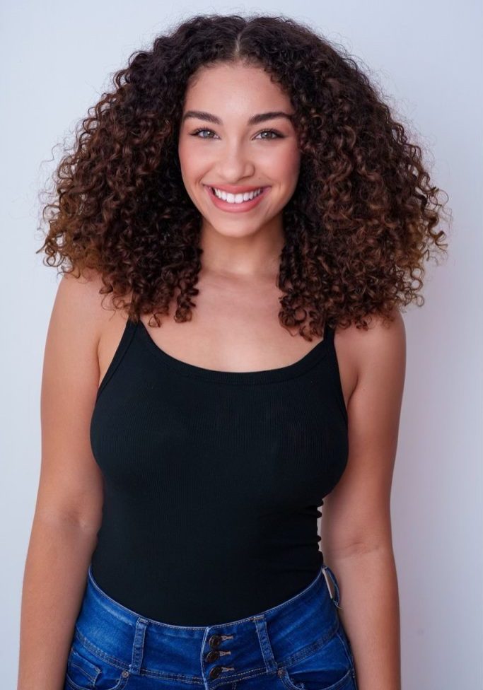 A woman with curly hair wearing a black tank top and blue jeans stands against a plain white background, smiling at the camera.