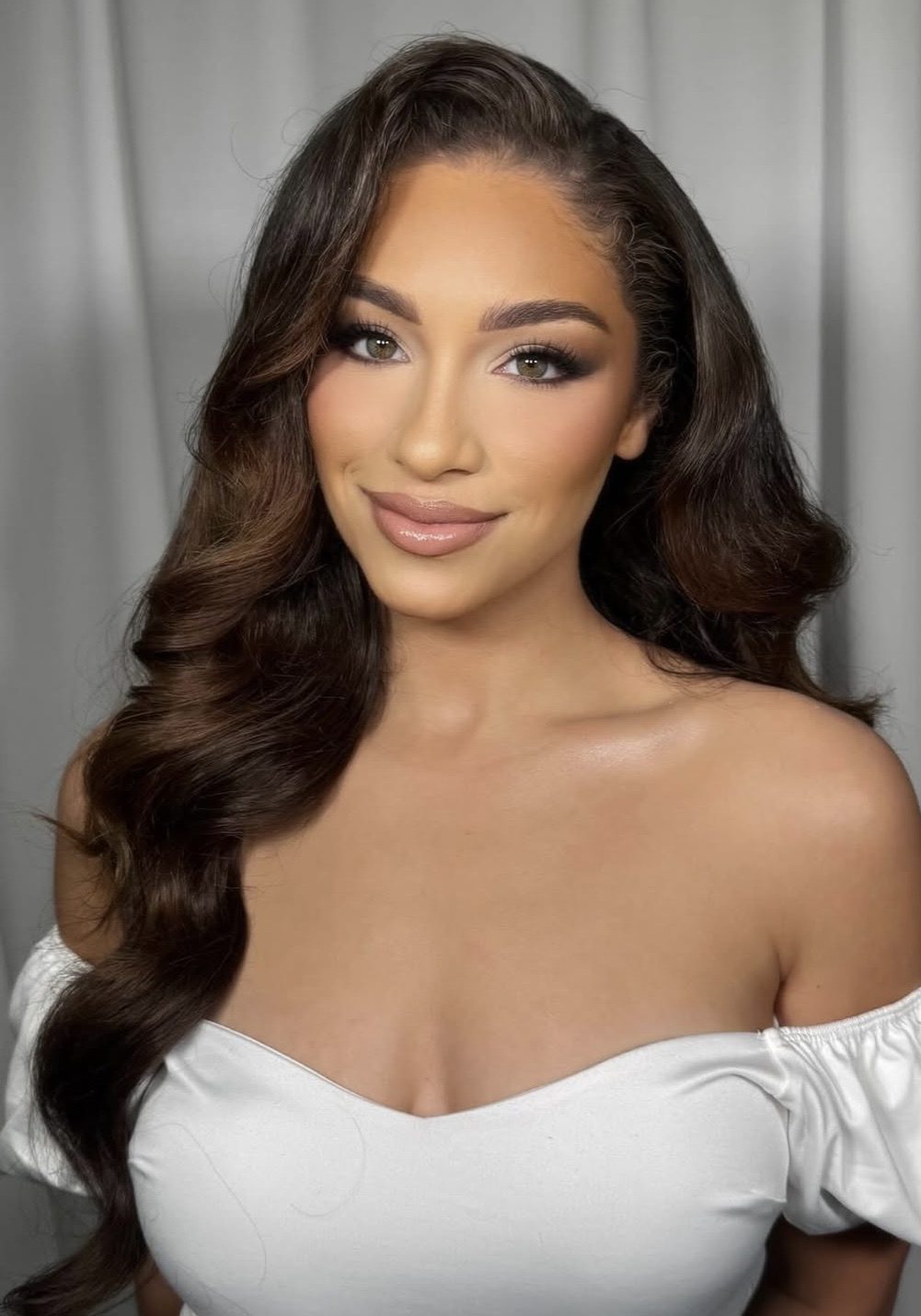 A woman with long wavy brown hair wearing an off-the-shoulder white top stands in front of a light gray curtain, smiling slightly at the camera.